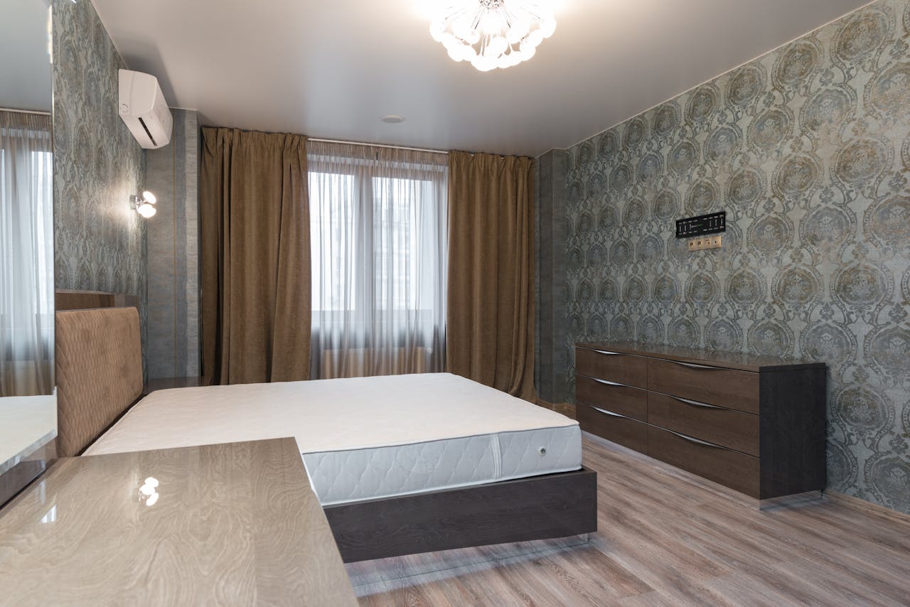 Interior of bedroom with air conditioning above bed placed near window with curtains and dresser
